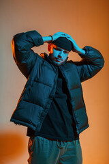 Cool stylish young handsome man in fashionable clothes with a winter down jacket puts on a hat and poses in a creative studio with orange and neon light