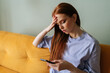 © dikushin - Closeup of upset unhappy young woman lady with redhaired in comfy homewear sitting on couch, using phone at home, looking at gadget screen and touching head, thinking about problem, reading bad news.