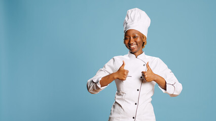 Wall Mural - Excited positive woman giving thumbs up on camera, earing kitchen apron and expressing like or approval symbol. Professional restaurant chef showing agreement gesture and success.