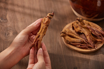 A red ginseng root in female hands on a matte brown wooden background. Red ginseng roots are placed on a plate next to the pot of decoction.
