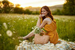 © VICHIZH - happy redhead woman sitting in a field of daisies on a plaid during sunset and smiling at the camera holding a flower in her hand