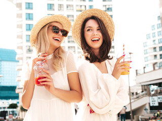 Wall Mural - Two young beautiful smiling hipster female in trendy summer white dress and straw hats. Sexy carefree women holding and drinking fresh vegetable cocktail smoothie drink in plastic cup with straw