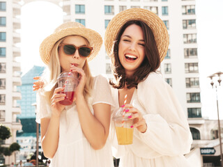 Wall Mural - Two young beautiful smiling hipster female in trendy summer white dress and straw hats. Sexy carefree women holding and drinking fresh vegetable cocktail smoothie drink in plastic cup with straw
