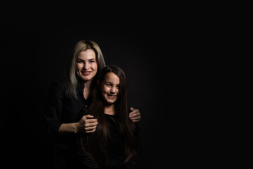 Wall Mural - Young mother and her little daughter are looking at camera and smiling, sitting against black wall