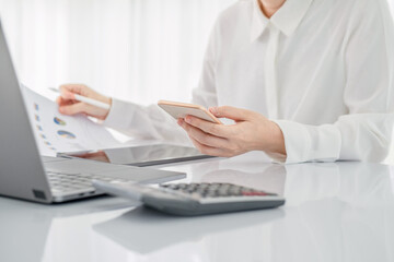 Asian businesswoman in white formal clothes writing note in tablet and checking smart phone. Business woman busy working with documents and tablet  in workstation. close up clean image.