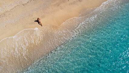 Wall Mural - Aerial view of beautiful happy woman in swimsuit sitting on sand in the shallow sea water, enjoying beach and turquoise ocean wave. Tropical sea in summer season on Egremni beach on Lefkada island.