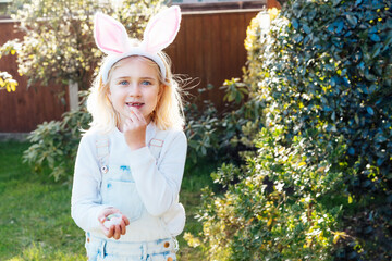 Little girl wearing bunny ears holding opened container with Easter chocolate egg sweets and eating candies after traditional egg hunting in garden. Kid having fun on Easter holiday. Selective focus.