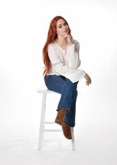 Wall Mural - full length portrait of beautiful woman model with long red hair, wearing casual outfit white blouse  top and denim jeans, isolated on white studio background. Relaxed seated pose, sitting on chair wi