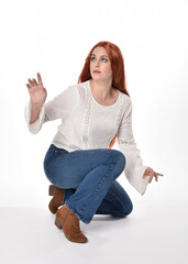 Canvas Print - full length portrait of beautiful woman model with long red hair, wearing casual outfit white blouse  top and denim jeans, isolated on white studio background. Relaxed seated pose, sitting on floor wi