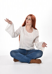 Wall Mural - full length portrait of beautiful woman model with long red hair, wearing casual outfit white blouse  top and denim jeans, isolated on white studio background. Relaxed seated pose, sitting on floor wi