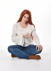 Wall Mural - full length portrait of beautiful woman model with long red hair, wearing casual outfit white blouse  top and denim jeans, isolated on white studio background. Relaxed seated pose, sitting on floor wi