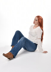 Wall Mural - full length portrait of beautiful woman model with long red hair, wearing casual outfit white blouse  top and denim jeans, isolated on white studio background. Relaxed seated pose, sitting on floor wi