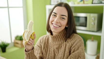 Wall Mural - Young beautiful hispanic woman holding banana sitting on table at kitchen