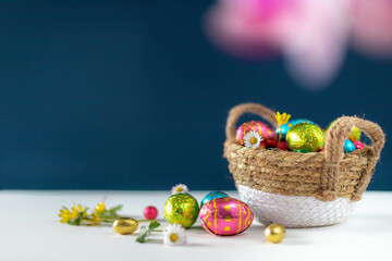 Wall Mural - Easter eggs in basket with blue background on white table. Studio lighting colorful easter decorated eggs for egg hunt with wicker basket and flowers. Banner for web and composition. Wishes card