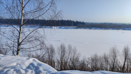 Wall Mural - Winter landscape with trees on a cliff and a view from a height of a frozen river or a field with snow on a cold sunny day with a blue sky