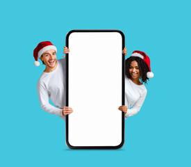 Poster - Christmas Offer. Happy Multiethnic Couple In Santa Hats Standing Behind Blank Smartphone