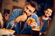 Young happy man eats burger and has fun with friends in pub.