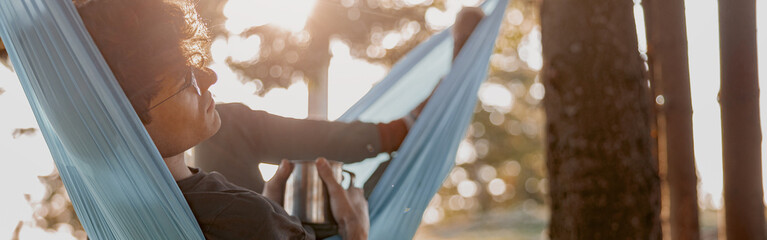 Wall Mural - Handsome young man in glasses lying in hammock and relaxing with coffee. Forest travel.