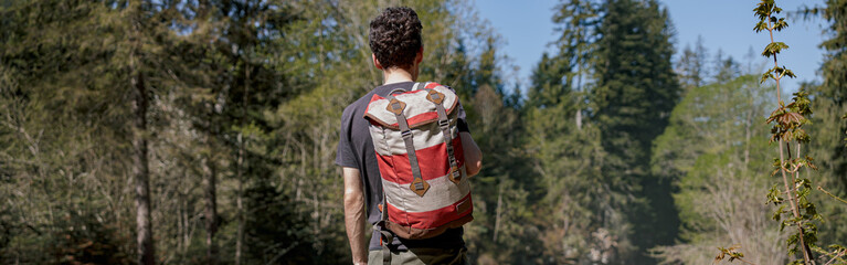 Wall Mural - Back view on male tourist with backpack standing in forest on cliff. Trekking travel. Tourism.