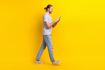 Sticker - Full size photo of handsome cheerful man gray t-shirt denim pants go empty space look at smartphone isolated on yellow color background