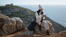 Romantic Girl Reading Book On Rocky Mountain. Serene Tourist Resting Ocean Cliff