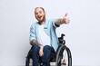 A man in a wheelchair looks at the camera shows a thumbs up, happiness, with tattoos on his hands sits on a gray studio background, health concept man with disabilities
