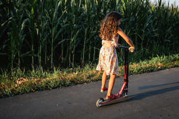 Wall Mural - girl in flower dress riding red scooter on a country road next to corn field