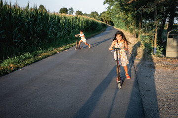 Wall Mural - two children riding red scooters on a country road next to corn field