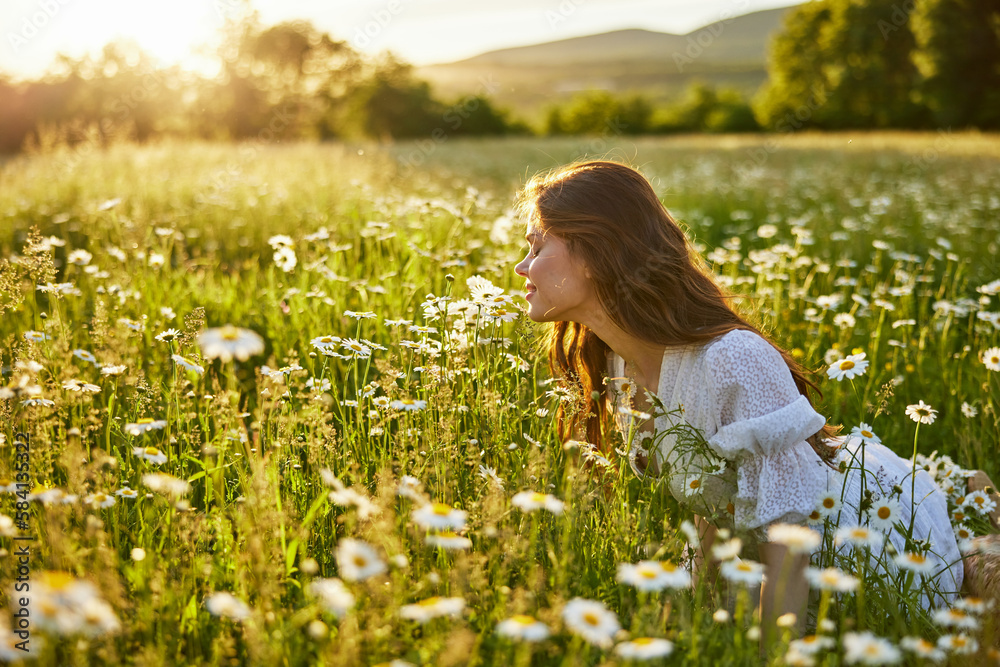 a beautiful woman in a light dress sits in a field of daisies against the backdrop of the setting sun and inhales their fragrance