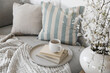 © tabitazn - Spring interior still life. Cup of coffee, tea on pile of books. Round beige table. Blossoming cherry plum tree branches in ceramic vase. Cozy linen sofa, cushions. Blurred background. Home decor. Top