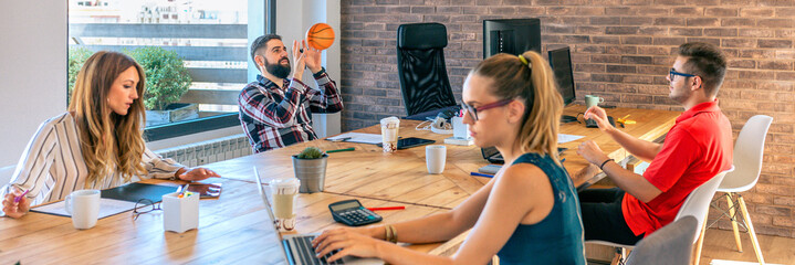 Wall Mural - Two young businessman having fun playing with a basket ball in coworking office while his female colleagues working. Selective focus on men in background. Relax and creativity time at office.