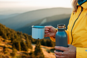 Refreshment after successfully climbed mountain summit. Female hiker drinking hot drink from travel mug and enjoying view at mountains during sunset