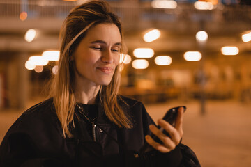 Wall Mural - Beautiful woman using smart phone in the city, smiling at camera at night, night city life. Girl try to call a taxi, girl chatting, texting in phone, look happy and smiling. Woman hold mobile.