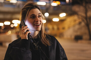 Wall Mural - Beautiful young woman talking on smartphone walking through night city street full of neon light. Portrait of gorgeous smiling female using mobile phone.