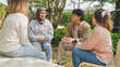 Small Asian group of people sit in a close circle and talk to a therapist in park. Smile people sharing story happy speak diverse people sitting in circle at group therapy session with psychologist.