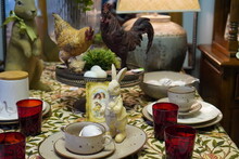 Easter Interior Of A Rabbit Figurine With Plates On The Table And Figurines Of A Chicken And A Cockerel On A Gray Wall Background