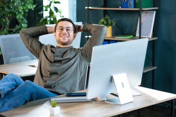 Relaxed confident business man taking break from work on computer for mind relief holding hands behind head thinking of success enjoying rest, good result of job done in office, no stress free concept