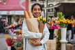 Young Latina woman smiling while buying yellow tulips and bread from a street vendor's stall, adding warmth to her day.