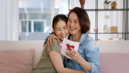 Wall Mural - May Mother's day young adult grown up child cuddle hug give flower gift box red heart card to mature middle aged mum. Love kiss care mom asia people sitting at home sofa happy smile enjoy family time.