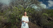 Smiling woman in white dress standing in a blooming almond orchard and throws up the white petals of flowers. Slow-motion.