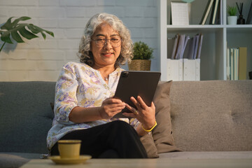 Pleasant modern senior woman relaxing on sofa at cozy home and using digital tablet. Elderly technology concept.