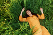 © VICHIZH - a relaxed red-haired woman enjoys summer lying in the tall green grass in a long orange dress smiling happily with her eyes closed