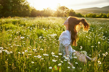A Red-haired Woman In A Light Dress Sits With Her Back To The Camera In A Field Of Daisies Straightening Her Hair With Her Hands Against The Backdrop Of The Setting Sun