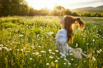 a red-haired woman in a light dress sits with her back to the camera in a field of daisies straightening her hair with her hands against the backdrop of the setting sun