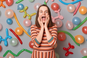 Wall Mural - Photo of extremely joyful woman with brown hair wearing striped dress standing around multicolored inflated balloons, yelling with happiness, being amazed, touching her face.