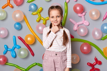 Wall Mural - Image of cute charming smiling little girl with braids wearing casual clothing posing isolated over gray background with balloons, holding her chin, showing thumb up.