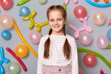 Wall Mural - Portrait of smiling delighted adorable little girl with braids wearing casual clothing posing isolated over gray background with balloons, looking at camera with happy face, celebrating birthday.