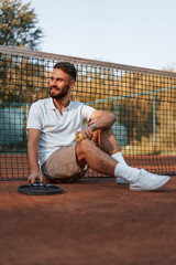 Wall Mural - Sitting on the ground near the net. Racket is lying down. Young man is on the tennis court at sunny daytime