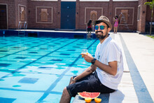Young Beautiful Indian Man Sitting On The Pool Edge With Feet In Water And Holding Glass Of Orange Juice In Hot Sunny Day. Guy Relaxing Outdoors By Swimming Pool. Summer Holiday And Vacation Concept.
