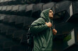 A man in a green rain jacket standing in front of a futuristic dark building in heavy rain wearing a backpack.